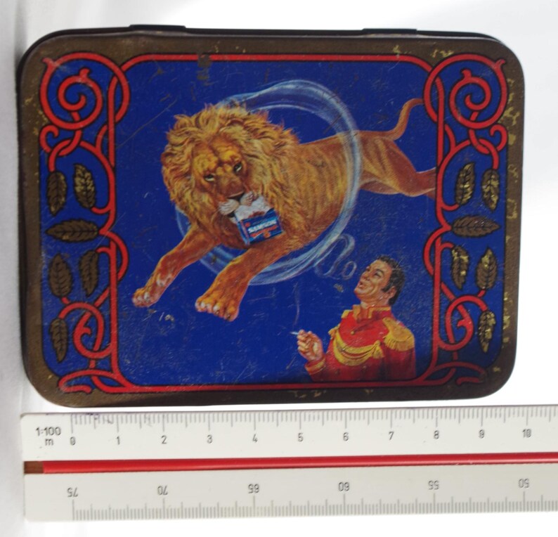 May include: A vintage metal tin with a blue background featuring a lion jumping through a smoke ring. A man in a red and gold uniform is looking up at the lion. The tin has a gold border with a red and gold design.