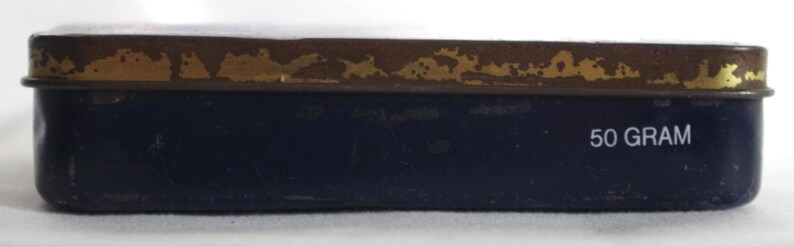 May include: A blue metal tin with a gold trim and the words "50 GRAM" printed on the bottom.
