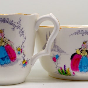 May include: A white porcelain creamer and sugar bowl set with a hand-painted design of a woman in a pink and blue dress. The design is surrounded by floral details and a gray vine.
