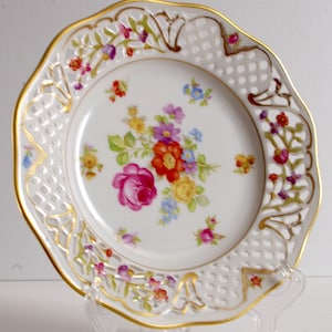 May include: A white porcelain plate with a gold rim and intricate cut-out design. The plate features a floral pattern with pink, red, yellow, and blue flowers.