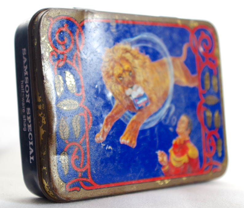 May include: Vintage tin box with a blue background featuring a lion performing a circus act. The text "SAMSON SPECIAL half-ware shag" is printed on the side of the box.