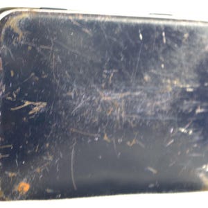 May include: A blue metal tin with a worn, scratched surface. The tin has a rectangular shape and is slightly dented.