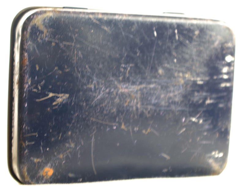 May include: A blue metal tin with a worn, scratched surface. The tin has a rectangular shape and is slightly dented.