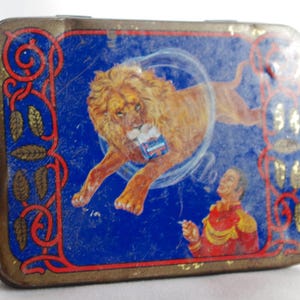 May include: Vintage tin box with a blue background featuring a lion jumping through a hoop. The lion is brown and has a white mane. A man in a red and gold uniform is standing on the right side of the box, holding a cigarette. The box has a gold border with a decorative pattern.