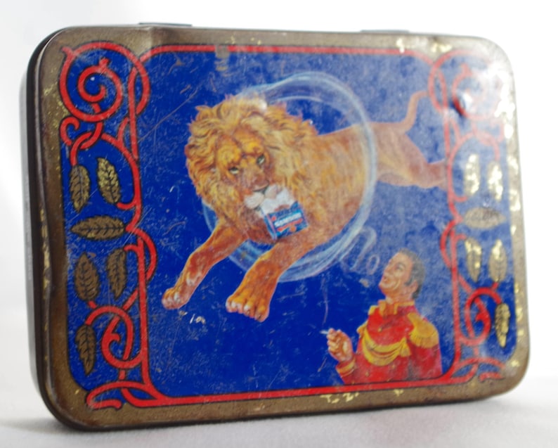 May include: Vintage tin box with a blue background featuring a lion jumping through a hoop. The lion is brown and has a white mane. A man in a red and gold uniform is standing on the right side of the box, holding a cigarette. The box has a gold border with a decorative pattern.