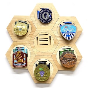 May include: A hexagonal wooden medal display with six slots for medals. The display is made of light brown wood and has a natural finish. The slots are arranged in a honeycomb pattern. There are six medals in the slots, each with a different design. The medals are all metal and have a variety of colors, including gold, silver, bronze, and blue.