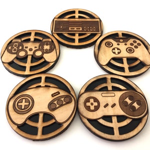 May include: Five wooden coasters with a gaming theme. Each coaster features a different game controller design, laser-cut into the wood. The coasters have a light brown color with a dark brown border, and are arranged in a circular pattern.