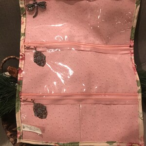 May include: A pink fabric jewelry roll with three clear zippered compartments. The roll has a floral print and the text "Heather Loft" is visible on the bottom right corner.