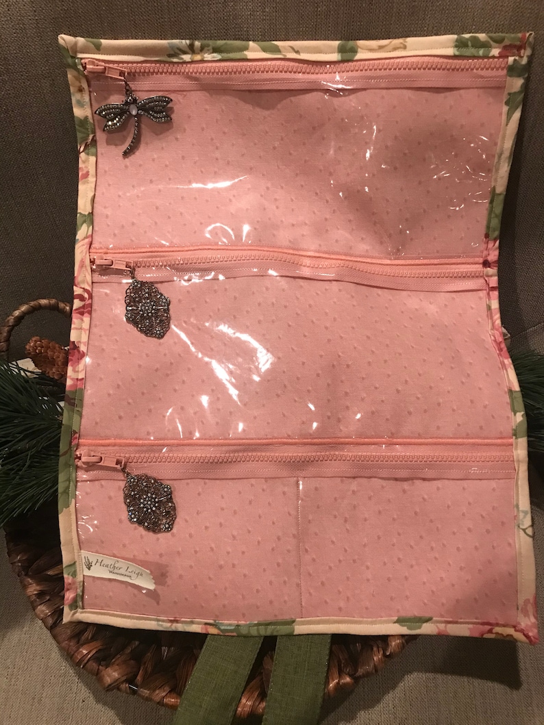 May include: A pink fabric jewelry roll with three clear zippered compartments. The roll has a floral print and the text "Heather Loft" is visible on the bottom right corner.