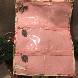 May include: A pink fabric jewelry roll with three clear zippered compartments. The roll has a floral print and a label that reads "Heather Loops".