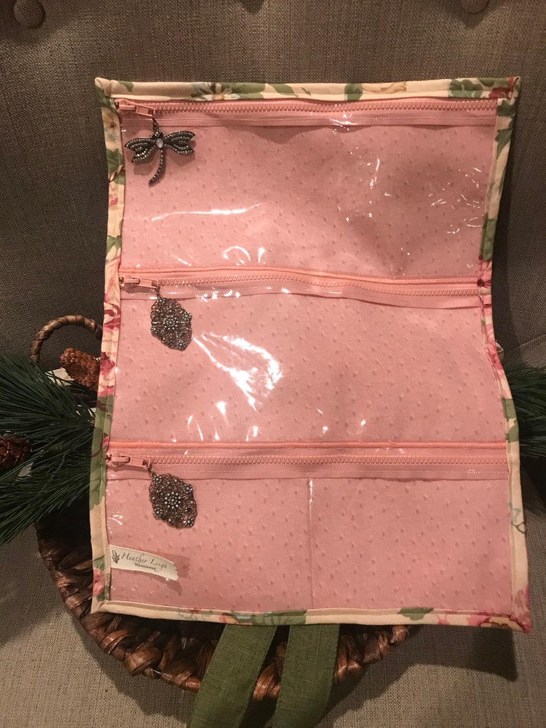 May include: A pink fabric jewelry roll with three clear zippered compartments. The roll has a floral print and a label that reads "Heather Loops".
