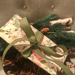 May include: A floral fabric gift bag with a green ribbon tied around it. The bag is sitting in a brown wicker basket.