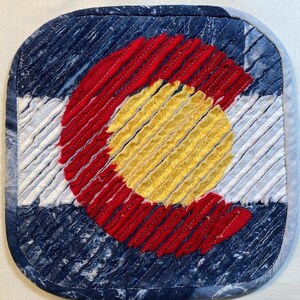 May include: A square pot holder featuring the Colorado state flag design. The flag is made of denim fabric and features a red, white and yellow circle with a white 'C' in the centre. The pot holder is quilted with red stitching.