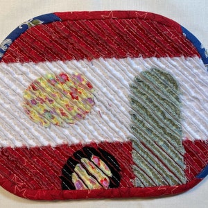 May include: Oval quilted fabric item in red, white, and blue. Features a red and white striped pattern, a floral-patterned circle, and a green and white striped shape. The edges are trimmed with a blue floral fabric, creating a decorative piece.