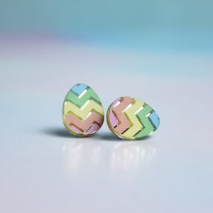 May include: A pair of pastel coloured egg-shaped earrings with a chevron pattern. The earrings are made of acrylic and have a gold-coloured backing.
