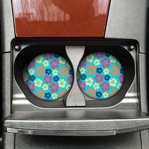 May include: Two round car coasters with a tropical floral pattern in shades of pink, yellow, blue, and green on a turquoise background. The coasters are in the cup holders of a car.