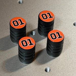 May include: Four cylindrical valve stem caps with a black knurled metal texture. Each cap features a circular orange top insert with the number 01 printed in a bold black font with a white outline, mimicking a classic racing car design element.
