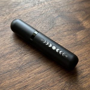 May include: A cylindrical matte black atomizer bottle featuring a white graphic print of the lunar cycle phases. The design displays a sequence of moon shapes, including crescents and a full circle, arranged in a horizontal line along the body.