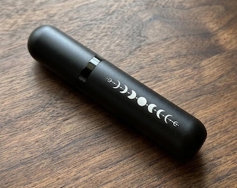 May include: A cylindrical matte black atomiser bottle featuring a white graphic print of the lunar cycle phases. The design displays a sequence of moon shapes, including crescents and a full circle, arranged in a horizontal line along the body.