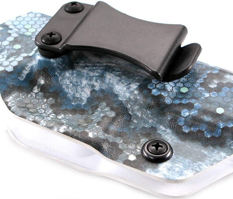 Pu&ograve; includere: Primo piano di una fondina con clip e viti nere. La fondina presenta un motivo esagonale blu, grigio e bianco. Il testo "Custom KYDEX Dig Camo Holsters" &egrave; visibile.