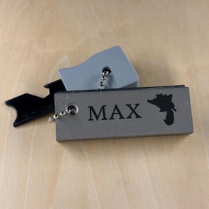 May include: A gray rectangular tag with the name "MAX" printed in black, along with a black silhouette of a state. A small black tool and a gray tag are also present. All items are attached to a ball chain.