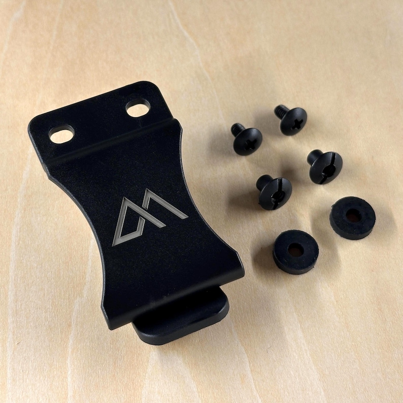 Pu&ograve; includere: Una staffa in metallo nero con un design geometrico e due fori per viti. La staffa &egrave; accompagnata da quattro viti nere e due rondelle in gomma nere. Gli elementi sono esposti su una superficie di legno chiaro.