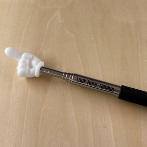May include: A white hand-shaped pointer with a thumbs-up gesture, attached to a silver metal telescoping pole. The text "Look Here, Genius" is printed on the pole. The handle is black.