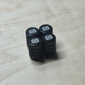 May include: Four black tire valve stem caps with a white "S" logo. The caps are cylindrical with a ridged design and are arranged in a square formation on a light wood surface. The caps are likely for a car or truck.