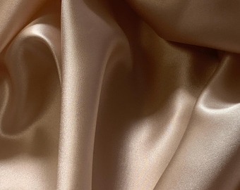 Puede incluir: Primer plano de una tela lujosa y fluida en un cálido color champán. El material tiene una textura suave y sedosa con sutiles reflejos y sombras, creando una sensación de profundidad y movimiento. La tela se pliega en suaves pliegues.