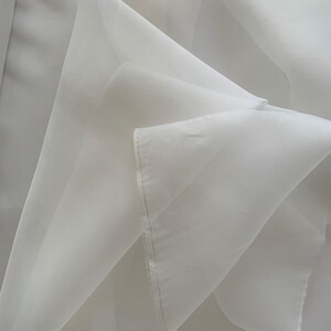 Könnte beinhalten: Nahaufnahme eines weißen, transparenten Stoffes mit zarter, fließender Textur. Der Stoff ist gefaltet und geschichtet, wodurch weiche Schatten und Glanzlichter entstehen. Die Kanten sind sauber mit einer feinen Naht genäht, was auf ein fertiges Kleidungsstück oder Textil hindeutet.