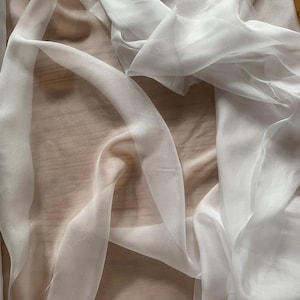 Puede incluir: Tela blanca transparente drapeada sobre una superficie marrón claro. La tela es ligera y fluida, con pliegues suaves y una calidad translúcida. El material parece ser seda o un textil delicado similar, ideal para ropa o decoración.