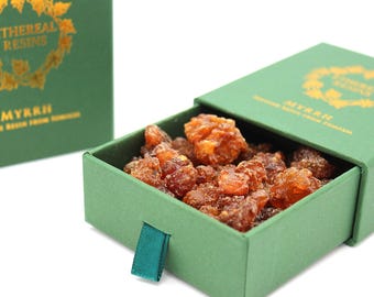 May include: A green sliding drawer box containing amber-colored myrrh resin pieces. The box features gold text reading ETHEREAL RESINS and MYRRH, with a decorative gold leaf wreath design. A small green fabric pull tab is attached to the front.
