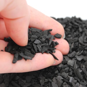 May include: A collection of small, irregular, matte black charcoal chips. The pieces are fragmented, porous, and dark in colour, with varying shapes and sizes ranging from tiny granules to slightly larger, splintered shards of carbonised material.