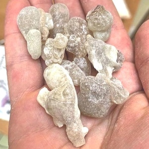 Puede incluir: Primer plano de un puñado de piezas de resina de color claro y forma irregular. La resina parece translúcida con una textura ligeramente granulada. Las piezas varían en tamaño y forma, algunas se asemejan a pequeñas formas redondeadas. El color general es blanquecino pálido.