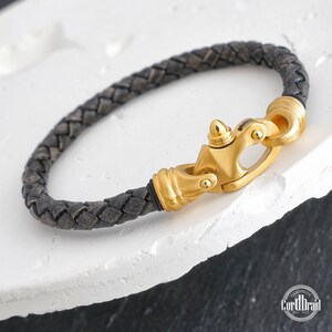 Puede incluir: Un brazalete trenzado de cuero negro con un cierre dorado. El cierre tiene un diseño único con una parte superior puntiaguda y una pequeña bola a cada lado. El brazalete está hecho a mano y presenta el texto "CordDraid est. 2015".