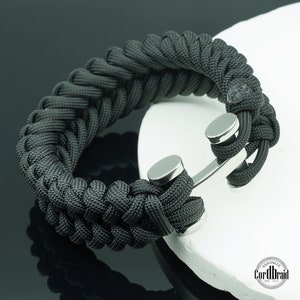 Könnte beinhalten: Schwarzes Paracord-Armband mit einem silbernen Ankerverschluss. Das Armband ist handgefertigt und hat ein gewebtes Design.