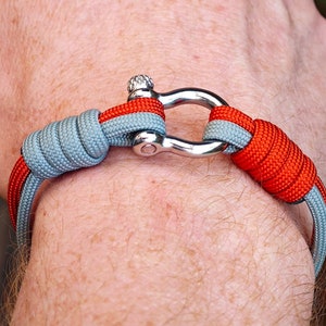 Könnte beinhalten: Nautisch inspiriertes Armband mit Schäkelverschluss aus Edelstahl. Das Armband besteht aus grauem und rotem Seil, das um das Handgelenk gewickelt ist. Ein stilvolles Accessoire, ca. 18 cm Umfang.