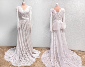 May include: A white lace wedding dress with long sleeves and a flowing skirt. The dress has a fitted bodice with a V-neckline and a belt at the waist. The skirt is made of layers of delicate lace and has a train.