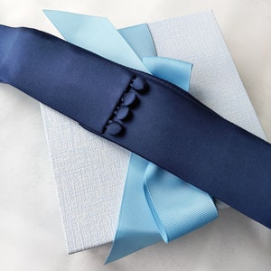 Può includere: Una fascia di raso blu navy con quattro piccoli bottoni coordinati è drappeggiata su una scatola strutturata azzurra, accentuata da un nastro azzurro. La fascia è il punto focale, suggerendo un accessorio formale.