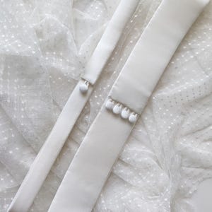 May include: Two white satin sashes with white pearl-like embellishments on the ends. The sashes are lying on a white tulle fabric with small white polka dots.