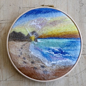Op de afbeelding: Een rond naaldkunstwerk dat een zonsondergang over een strand met golven die op de kust breken afbeeldt. Het strand is zanderig met voetafdrukken die naar het water leiden. De lucht is een gradiënt van blauw, geel en oranje.