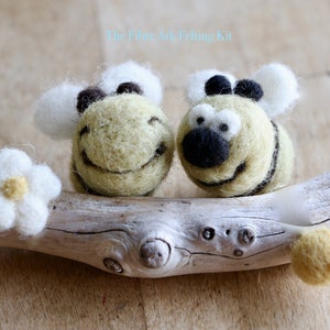 Puede incluir: Dos abejas sonrientes de fieltro hechas a mano con rayas negras y amarillas y alas blancas descansan sobre un trozo de madera flotante. Una flor de fieltro blanco y una bola de fieltro amarillo también están en la madera flotante.