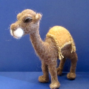 May include: A brown and tan felt camel figurine with a saddle. The camel has a long neck and a hump on its back. The saddle is made of felt and has a brown rope design.