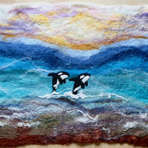 May include: A needle felted wall art piece depicting two orcas swimming in a blue ocean with a sunset sky above. The ocean is layered with shades of blue and the sky is layered with shades of pink, purple, and yellow.