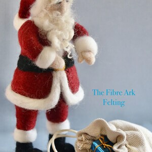 Könnte beinhalten: Eine Filz-Weihnachtsmann-Puppe, die einen roten Anzug mit weißen Verzierungen und einen schwarzen Gürtel trägt. Er hält einen weißen Sack mit verpackten Geschenken. Der Text "The Fibre Ark Felting" ist im Hintergrund sichtbar.