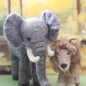 May include: A grey felt elephant and a brown felt lion stand side-by-side on a green surface. Both animals have a realistic appearance and are made with soft, fuzzy felt.