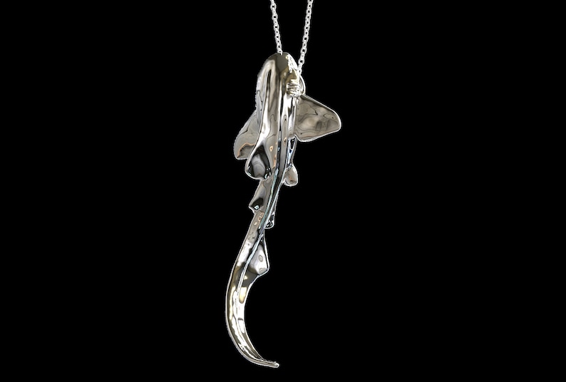 May include: A silver-colored pendant necklace featuring a detailed, three-dimensional sculpture of a shark. The shark is suspended from a silver chain.
