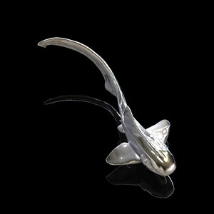 May include: A silver-colored shark sculpture with a long, curved tail. The sculpture is posed as if it is swimming, with its head tilted upwards. The sculpture is set against a black background.