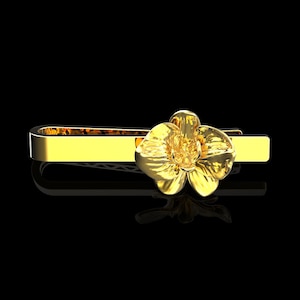 May include: A gold-colored tie clip featuring a detailed orchid flower design. The tie clip is rectangular with a floral accent, set against a black background. The clip is likely made of metal.