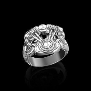 Può includere: Un anello d'argento con un design dettagliato di un motore di motocicletta. L'anello è in metallo e ha una finitura lucida.